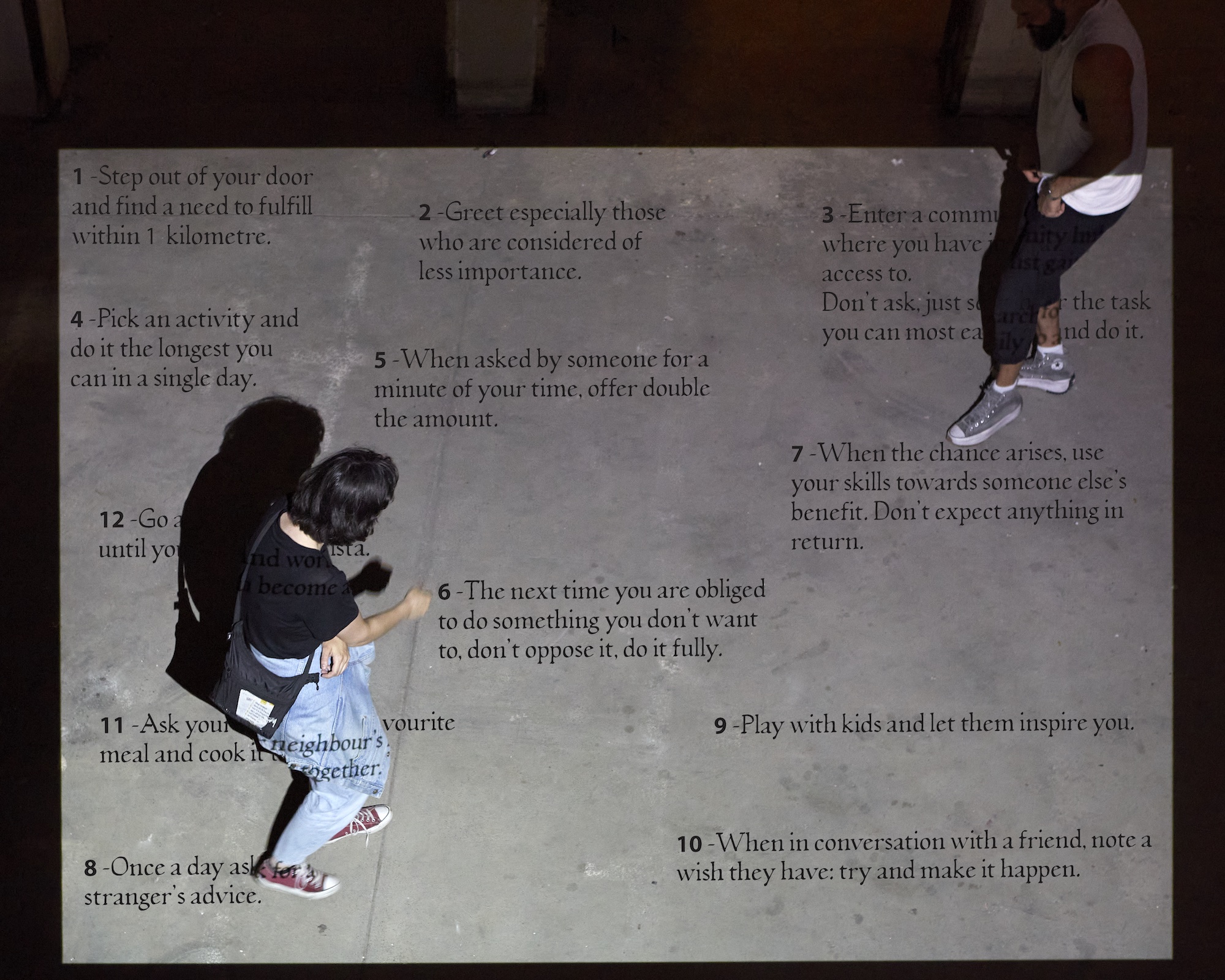 Overhead view of two people standing on a concrete floor. A large projection on the floor lists the guidelines of the project.